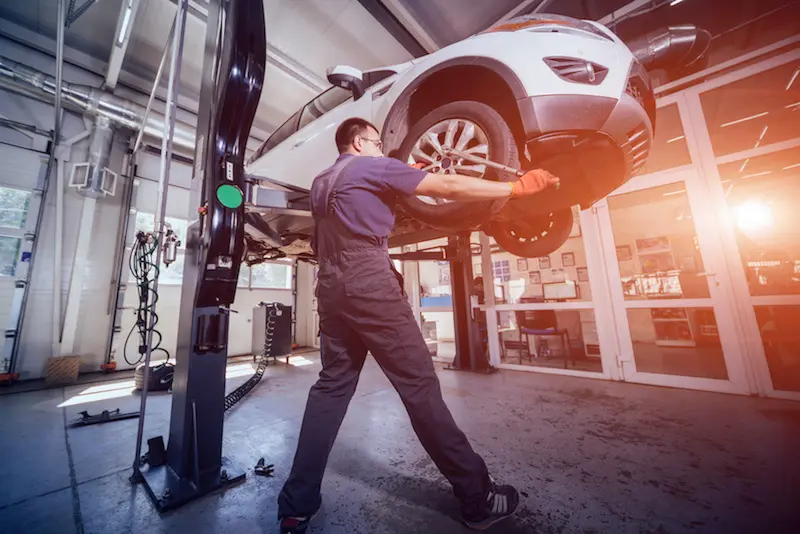 Car mechanic inspecting wheel and suspension detail of lifted automobile at repair service station