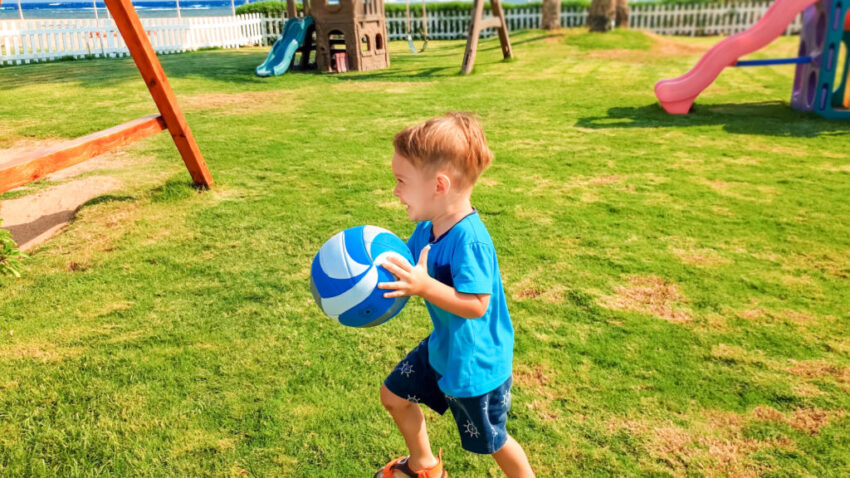 Portrait of happy smiling little boy holding ball in hands and running on grass at children playground