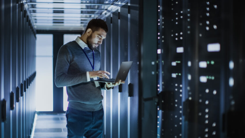 It,Technician,Works,On,Laptop,Next,To,A,Server,Cabinet